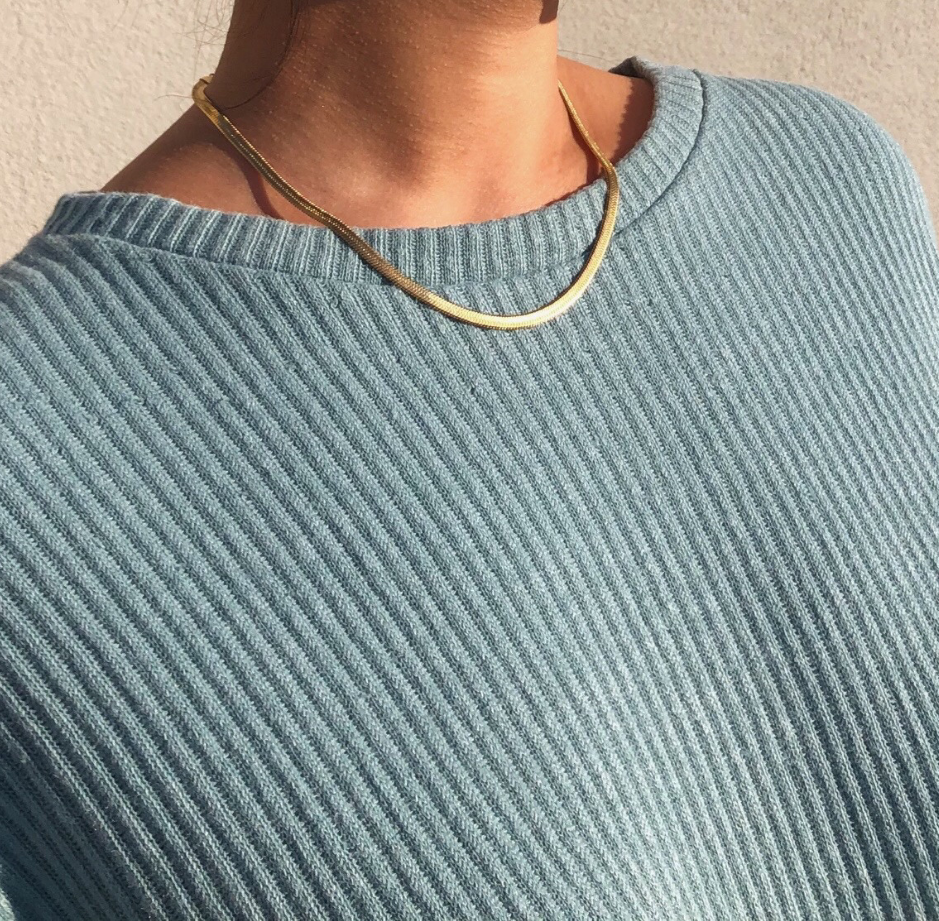 A gold herringbone necklace laying across a blue knit sweater demonstrating how to elevate a look with a simple dainty necklace.